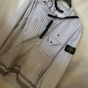 Stone Island rutig vindjacka med huva - Säljer en vit och svart rutig vindjacka från Stone Island med huva och dragkedja framtill. Jackan har klassisk patch på ärmen, bröstficka med knapp och stora loggan på ryggen. Tillverkad i polyester och har en loose passform, perfekt för dig som gillar streetwear vibes.