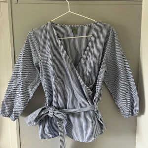 Randig omlottblus med knytband - Skitnygg blåvit randig omlottblus från Lindex med trekvartsärmar och knytband i midjan. Blusen har v-ringning och slutsåld på hemsidan🤗