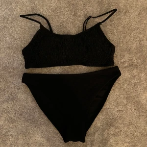 Svart bikini med smockad topp - Snygg svart bikini med smala axelband och smockad topp som ger en cool textur. Bikinitrosan har en klassisk, något hög midja och är tillverkad i ett stretchigt material som sitter skönt. Perfekt för stranden eller poolen i sommar!