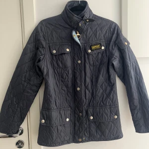 Mörkblå quiltad jacka från Barbour - Quiltad mörkblå jacka från Barbour International med guldiga detaljer, hög krage med spänne och flera fickor framtill. Klassisk look med Barbour-logga på bröstet och dragkedja med tryckknappar. Perfekt för dig som gillar stilren och tidlös design. Mycket bra skick.