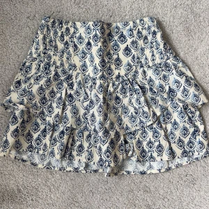 Vit och blå mönstrad kortkjol - Superfin kortkjol i vitt med blått mönster och volangdetaljer. Kjolen har resår i midjan för skön passform och är gjord i ett lätt bomullstyg. Perfekt för sommaren!! 😍😍Inga defekter då den ej är använd. 