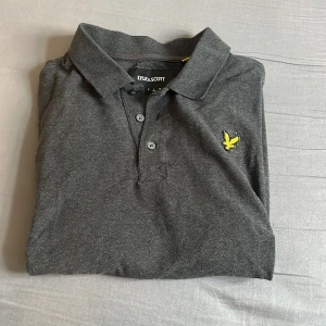 Mörkgrå pikétröja från Lyle & Scott - Snygg mörkgrå pikétröja från Lyle & Scott med klassisk krage och tre knappar framtill. Tröjan har det ikoniska gula fågel-loggan broderad på bröstet. Tillverkad i mjuk bomull och passar perfekt till en avslappnad men stilren look. Hör av er vid minsta lilla fråga. 