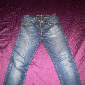 Blåa jeans från Nudie Jeans Co - Säljer ett par klassiska blåa jeans från Nudie Jeans Co med snygga slitningar och kontrastsömmar. Byxorna har fem fickor, orange broderi på bakfickorna och läderpatch i midjan. Perfekta för dig som gillar en tidlös och avslappnad stil. W31/L32. De är i slimfit passform och passar klockrent.