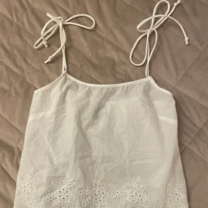 Vit broderad linnetopp från H&M - Supersöt vit linnetopp från H&M med tunna axelband som knyts och broderade blommor längst ner. Perfekt för varma dagar och ger en fräsch och somrig vibe. Lätt och luftig känsla tack vare bomullsmaterialet💗💗