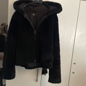 Svart pälsjacka med huva från ONLY - Mysig svart pälsjacka från ONLY med huva och dragkedja framtill. Jackan har en clean look och är perfekt för dig som vill ha en stilren vibe under kyliga dagar. Passformen är normal och materialet känns mjukt och fluffigt.