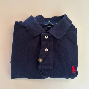 Marinblå pikétröja från Ralph Lauren - Klassisk marinblå pikétröja från Ralph Lauren med röd broderad logga på bröstet. Tröjan har korta ärmar, krage och knäppning med tre knappar. Tillverkad i bomull och har en snygg custom fit-passform. Perfekt för en stilren och avslappnad look.