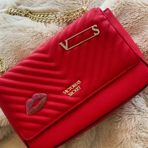 Röd quiltad axelväska Victoria's Secret - Säljer en snygg röd axelväska från Victoria's Secret med quiltad design, guldfärgad kedja och coola detaljer som VS-logga och glittrande läppar. Väskan har en stilren form och är gjord i syntetmaterial. Perfekt för dig som vill sticka ut med en färgstark accessoar.