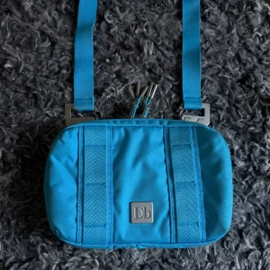 Db Crossbody Bag - Snygg och smidig axelväska från Db / Douchebag i en klar isblå färg. Väskan har justerbar rem, flera fack inuti och en silverfärgad logga framtill. Tillverkad i slitstark polyester och perfekt för att bära det viktigaste på stan eller resan. Mycket bra kvalité. Nypris 1350kr. Bra skick, knappt använd.