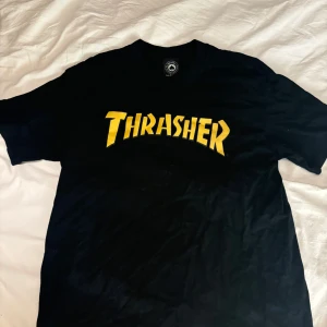 Svart Thrasher t-shirt med gul logga - Svart t-shirt från Thrasher med ikonisk gul logga tryckt över bröstet. Klassisk passform med rund hals och korta ärmar. Perfekt för dig som gillar streetwear och skatekultur. Materialet är mjuk bomull som känns skönt mot huden. Nyskick storlek medium 