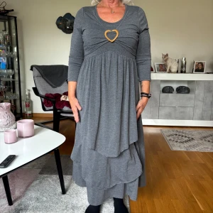 Grå långklänning med hjärtdetalj - Superfin grå långklänning med trekvartsärm och lager-på-lager kjol. Snygg hjärtformad detalj framtill och rundad halsringning. Klänningen har en avslappnad passform och är gjord i mjukt bomullsmaterial. Perfekt för dig som gillar en stilren och bekväm look.