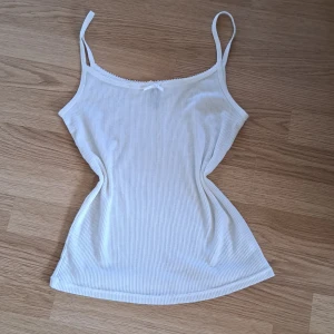 Vit ribbad linnetopp med rosett - Supersöt vit ribbad linnetopp med smala axelband och liten rosett framtill. Toppen har en feminin spetskant vid urringningen och är perfekt för varma dagar eller som lager under en skjorta. Enkel och stilren design som passar till allt. Aldrig använd. Lite genomskinlig💕