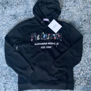 Alexander McQueen hoodie - Svart hoodie från Alexander McQueen med färgglad logga framtill. Endast testad och i sprillans nytt skick. Hör av er vid funderingar😊