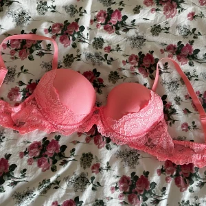Rosa spets-bh med vaddering - Snygg rosa bh med vadderade kupor och detaljer i spets. Axelbanden är justerbara och bh:n har en feminin look med blommig spets längs kanterna. Perfekt för dig som gillar färg och fina detaljer.