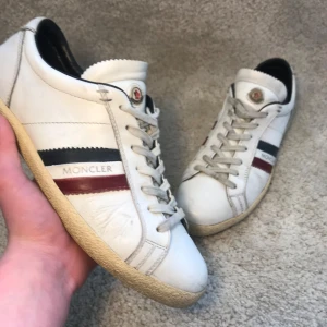 Vita sneakers från Moncler med ränder - Snygga vita sneakers från Moncler med klassiska blå och röda ränder på sidan och Moncler-logga på plösen. Skorna har snörning, rund tå och platt sula. Tillverkade i skinn för en lyxig känsla och passar perfekt till en clean streetwear-look.