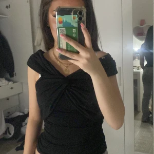 Svart omlott topp med twistad detalj - Säljer en svart omlott topp med snygg twistad detalj framtill. Toppen har offshoulder-design och sitter tight, perfekt för att lyfta din stil. Mjuk och stretchig känsla, passar till många olika outfits.