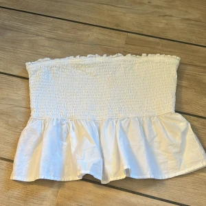 Vit bandeau tubtopp från Gina Tricot - Supersöt vit bandeau tubtopp från Gina Tricot med smockad överdel och volang nertill. Toppen är axelbandslös och har en lätt rynkad kant upptill för extra detalj. Perfekt för varma dagar och enkel att styla till jeans eller kjol. Den har inga defekter och är aldrig använd då den är för stor.