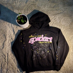 Svart Sp5der hoodie - Design och Utseende Hoodiens helsvarta bas gör att den rosa "Spider"-texten verkligen poppar. Texten ramas in av ett grått nätmönster som ger ett futuristiskt intryck, och färgglada stjärnor i gult, blått och rosa tillför en lekfull kontrast. Resultatet är en stilren men iögonfallande design.  Passform Jag testade storlek M (55 cm axelbredd, 65 cm längd). Passformen är avslappnad men ändå snyggt formad. Den sitter bra över axlarna och är bekväm att bära både till vardags och som statementplagg. 