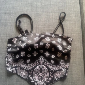 Svart bandana-linnetopp med paisleymönster - Snygg svart linnetopp med tunna axelband och paisleyinspirerat bandanamönster i vitt. Toppen har en spetsig nederkant och knytning i ryggen för extra cool detalj. Perfekt för dig som vill sticka ut med en trendig och edgy stil.