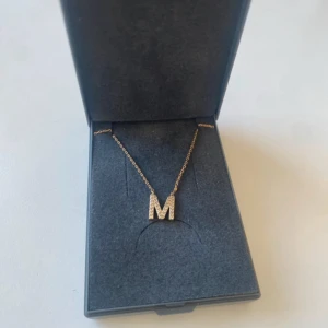 Guldigt halsband med bokstaven M - Snyggt halsband i guld med en glittrig bokstav M som hänge. Kedjan är tunn och elegant, och bokstaven är täckt av små gnistrande stenar som ger extra bling. Perfekt accessoar för dig som vill ha något personligt och trendigt.