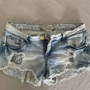 Jeansshorts med paljettdetaljer Simply Chic - Snygga ljusblå jeansshorts från Simply Chic med slitna detaljer och silverfärgade paljetter på framsidan. Rå kant nertill och klassisk femficksmodell. Perfekta för sommaren när du vill sticka ut lite extra.
