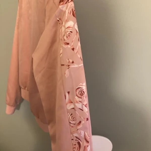 Rosa bomberjacka Puma Kenza XS - Snygg rosa bomberjacka från Puma x Kenza med blommiga detaljer på axlar och ärmar. Jackan har guldig Puma-logga framtill, ribbade muddar och klassisk bomberkrage. Perfekt för dig som vill sticka ut med en unik och trendig jacka.