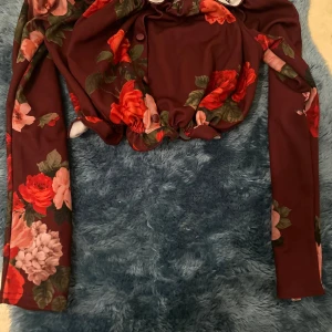 Blommig croppad bolerotopp New Look - Supersöt croppad bolerotopp från New Look i vinröd nyans med stora blommor i rött, rosa och grönt. Toppen har långa ärmar, knappar framtill och elastisk kant vid midjan. Perfekt för dig som vill sticka ut med en romantisk vibe.