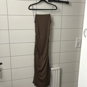 Brun långklänning med knutdetalj och slits - Säljer en brun långklänning med tunna axelband och snygg knutdetalj framtill som skapar en liten cut-out. Klänningen har en rak siluett och är tillverkad i ett glansigt, mjukt material som ger en lyxig känsla. Perfekt för dig som gillar minimalistisk och trendig stil.