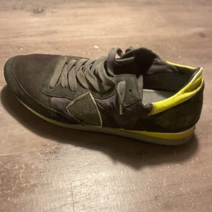 Philippe model  med gula detaljer - Snygga svarta sneakers med coola gula detaljer på hälen och sulan. Skorna har snörning, rund tå och platt sula. Materialmix av mocka och syntet ger en sportig vibe. Perfekta för dig som gillar streetstyle och vill sticka ut lite extra.