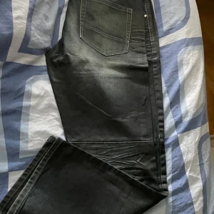 Svarta loose fit jeans KappAhl 1953 - Säljer ett par svarta jeans från KappAhl 1953 Denim Brand i loose fit. Jeansen har coola slitningar och markerade sömmar över benen, fem fickor och klassisk knappgylf. Perfekta för dig som gillar avslappnad stil och vill ha extra utrymme i benen.