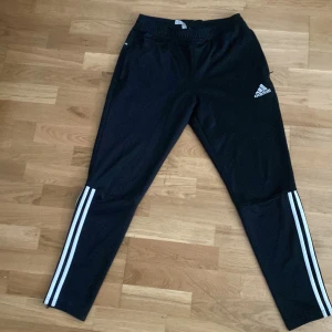 Svarta Adidas träningsbyxor - Svarta träningsbyxor från Adidas med klassiska vita ränder längs benen och logga på låret. Byxorna har smal passform och resår i midjan, perfekta för sport eller chill. Snygg och sportig look!