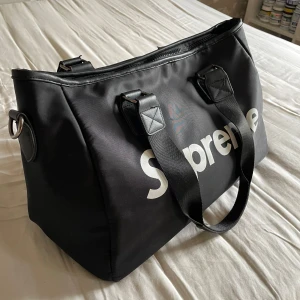 Supreme axelväska - Stor svart axelväska från Supreme med tydlig vit logga på framsidan. Väskan har dubbla breda handtag, dragkedja upptill och är tillverkad i slitstark polyester. Perfekt för dig som vill ha en streetstyle-vibe och behöver plats för allt du behöver under dagen.