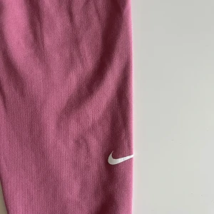 Rosa Nike Dri-FIT leggings - Snygga rosa leggings från Nike med Dri-FIT-teknologi. De har en hög midja och är perfekta för träning eller chill. Materialet är stretchigt och andas, med en vit Nike-logga på benet för extra stilpoäng.