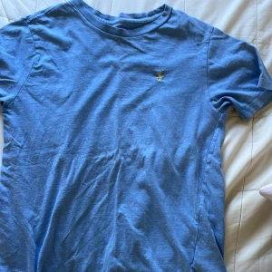 Blå t-shirt från Polo Ralph Lauren - Snygg blå t-shirt från Polo Ralph Lauren med klassisk passform och liten gul broderad logga på bröstet. Tillverkad i mjuk bomull och har rund halsringning samt korta ärmar. Perfekt för en avslappnad och stilren look.