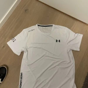 Vit Under Armour t-shirt - Snygg vit t-shirt från Under Armour med svart logga på bröstet och tryck på ärm och sida. Klassisk rund hals och korta ärmar, perfekt för träning eller chill. Tillverkad i ett lätt och snabbtorkande material som andas.