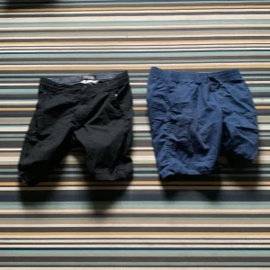 Svarta och blå shorts H&M & Lindex - Två par shorts i storlek 152, ett par svarta från H&M med knapp och dragkedja samt fickor, och ett par blå från Lindex med resår i midjan och sidfickor. Perfekta för sommardagar och chill häng. Båda är i mjuk bomull och har klassisk, enkel design.
