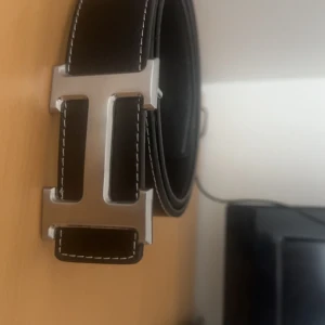 Svart skärp med silverfärgat H-spänne - Snyggt svart bälte i skinn med ett stort, silverfärgat H-format spänne. Bältet har vita kontrastsömmar längs kanterna som ger en clean och modern look. Perfekt accessoar för att lyfta din outfit med en exklusiv touch.