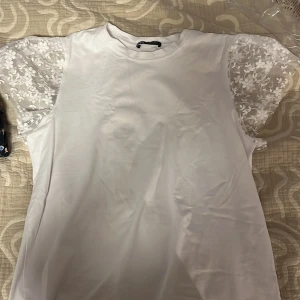 Vit blus med spetsärmar från Zara - Superfin vit blus från Zara med korta ärmar i transparent spets och blommigt broderi. Klassisk rund halsringning och rak passform. Perfekt för dig som vill ha en söt och stilren topp med det lilla extra i ärmdetaljerna.