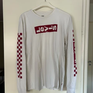 Vit långärmad Levi's tröja med tryck - Vit långärmad tröja från Levi's med rött logotryck på bröstet och schackrutigt mönster längs ärmarna. Tröjan har en klassisk passform och är gjord i mjuk bomull, perfekt för en avslappnad streetstyle-look. Nypris 999kr