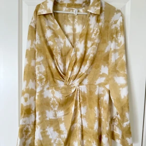 Beige och vit omlottklänning H&M - Superfin kort omlottklänning från H&M i beige och vitt batikmönster. Klänningen har långa ärmar, v-ringning och snygg twistad detalj framtill. Lätt och flowy känsla, perfekt för varmare dagar. Stängs med en liten hake vid bysten.
