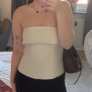 Beige bandeau tubtopp i linne - Säljer en stilren beige bandeau tubtopp i linne. Toppen är axelbandslös och har en snygg volangdetalj upptill samt en unik vikning baktill. Perfekt för dig som gillar minimalistisk och trendig stil. Oanvänd, sitter som en S