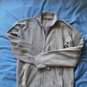 Beige ribbad kofta med dragkedja - Säljer en beige ribbad kofta från Dressmann med hel dragkedja framtill och ståkrage. Tröjan har långa ärmar och diskreta fickor på sidorna. Perfekt för lager-på-lager och chill stil. Passar dig som gillar enkel och stilren look.