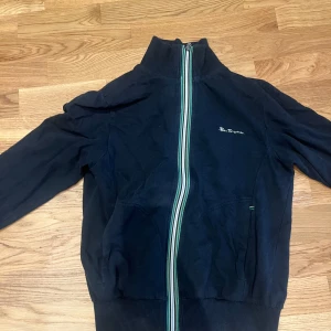 Mörkblå fullzip från Ben Sherman S - Mörkblå fullzip-tröja från Ben Sherman med hel dragkedja framtill och ribbade muddar. Jackan har en cool randig detalj längs dragkedjan i grönt och vitt samt diskret logga på bröstet. Perfekt för dig som gillar en stilren och sportig look.