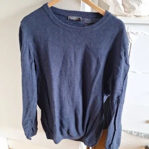 Mörkblå stickad tröja från Dressmann - En klassisk mörkblå stickad tröja från Dressmann i storlek L. Tröjan har rund halsringning, långa ärmar och ribbade muddar. Perfekt för lager-på-lager och enkel att matcha med jeans eller chinos.