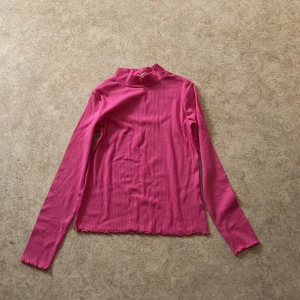 Rosa ribbad polotröja - Säljer en rosa ribbad polotröja med lång ärm och hög hals. Tröjan är figurnära och har en mjuk, stretchig känsla. Perfekt att styla med jeans eller kjol för en trendig look.