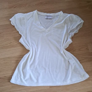 Vit blus med spetsärmar Holly & Whyte - Superfin vit blus från Holly & Whyte med v-ringning och korta ärmar i broderad spets. Luftig passform och mjukt bomullstyg gör den perfekt för varma dagar. Ärmarna har blommiga broderier som ger en söt och romantisk vibe💕