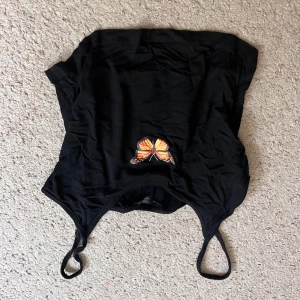 Svart linne  - Svart linne med smala axelband och en broderad orange fjäril framtill. Snygg croppad modell som passar perfekt till jeans eller kjol. Enkel men ändå unik detalj med fjärilen som sticker ut.