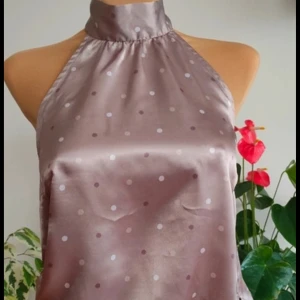 Prickig halterneck topp i satin - Snygg halterneck topp i glansig satin med lila och vita prickar. Toppen har hög hals med knyt i nacken och en lös passform som knyts i midjan. 