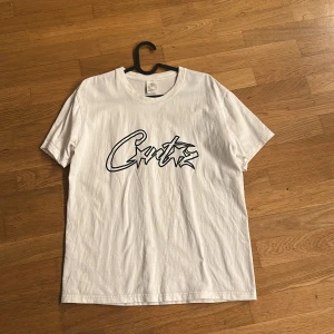 Vit Corteiz t-shirt med tryck - Vit t-shirt från Corteiz med svart grafiskt tryck på bröstet. Klassisk rund halsringning och korta ärmar. Perfekt för en chill och stilren look.