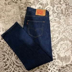 Levi's 501 mörkblå jeans. - Klassiska Levi's 501 jeans i mörkblå denim med raka ben och normal passform. Byxorna har fem fickor, gylf med knappar och den ikoniska läderpatchen bak i midjan. Perfekta för en tidlös och avslappnad look.