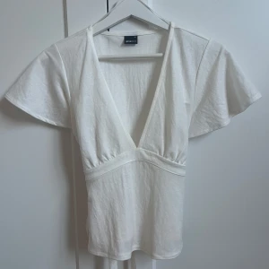 Vit v-ringad topp med knyt från Gina Tricot - Säljer en vit topp från Gina Tricot i storlek XS. Toppen har djup v-ringning, korta ärmar och knyt i midjan baktill. Perfekt för dig som gillar en enkel men snygg stil. Använd en gång så är precis som ny💗